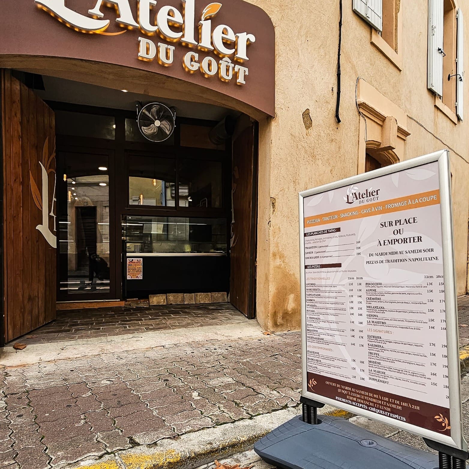 Vue de l'entrée de l'établissement "L'Atelier du Goût" avec un stop trottoir affichant le menu.