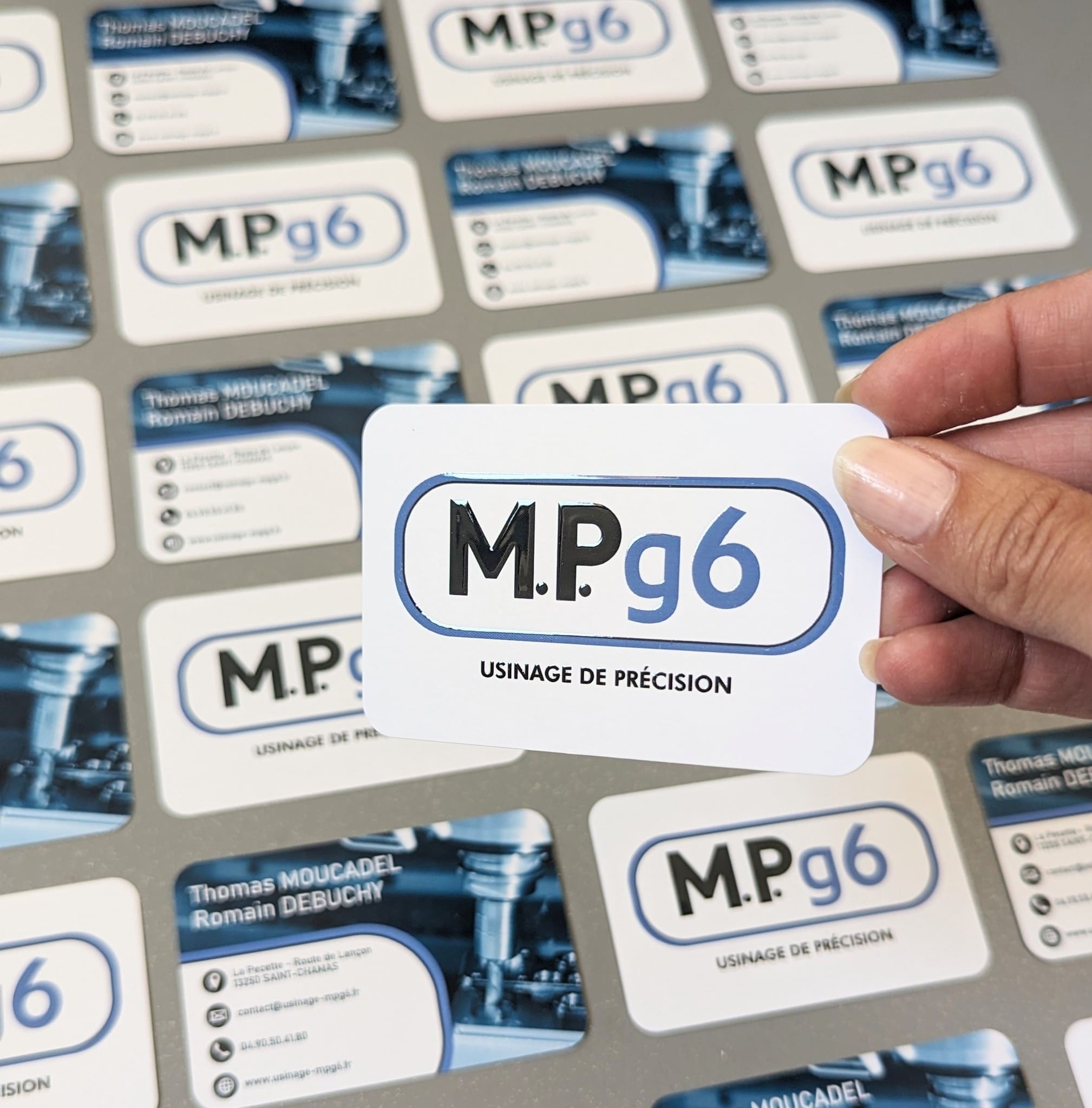 Main tenant une carte de visite MPg6 sur fond composé de cartes identiques.