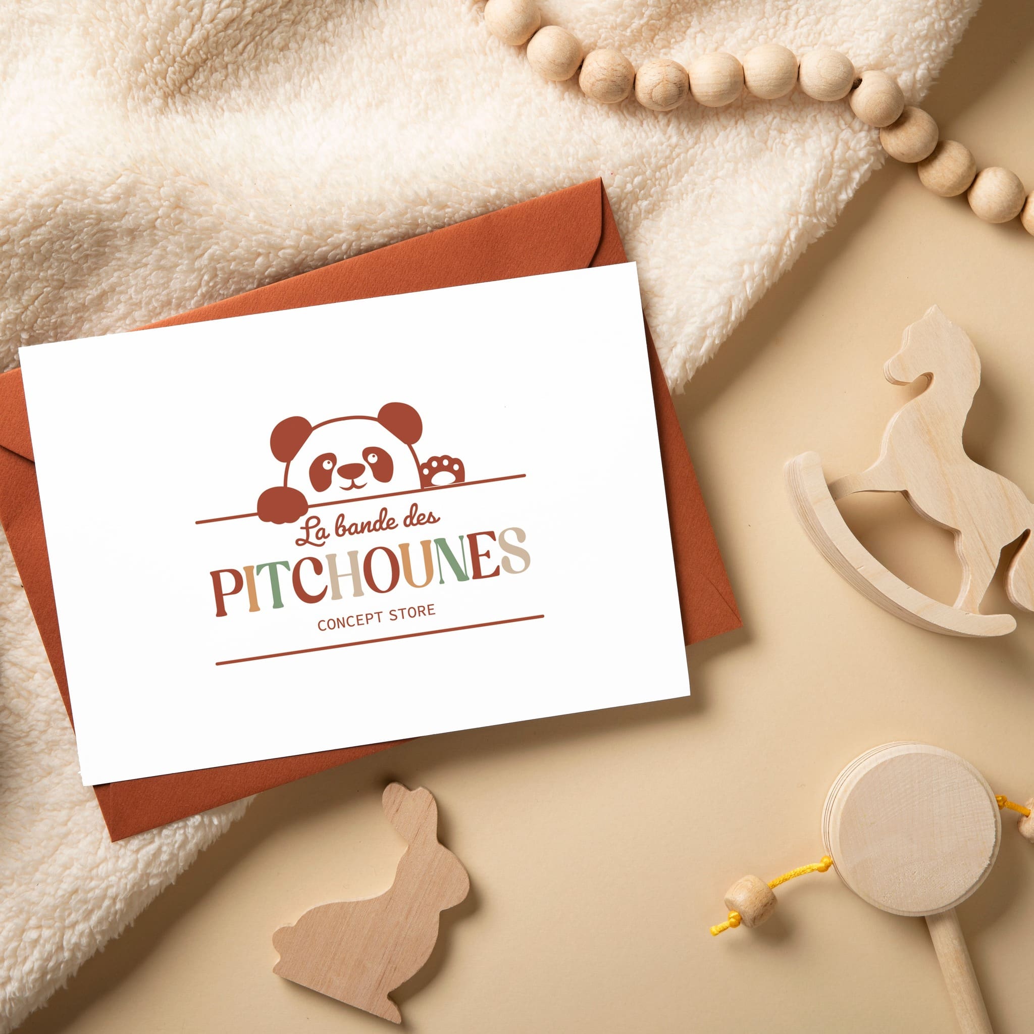 Carte de visite posée sur une enveloppe, entourée de jouets en bois et d’un plaid beige, avec le logo "La bande des Pitchounes - Concept Store".
