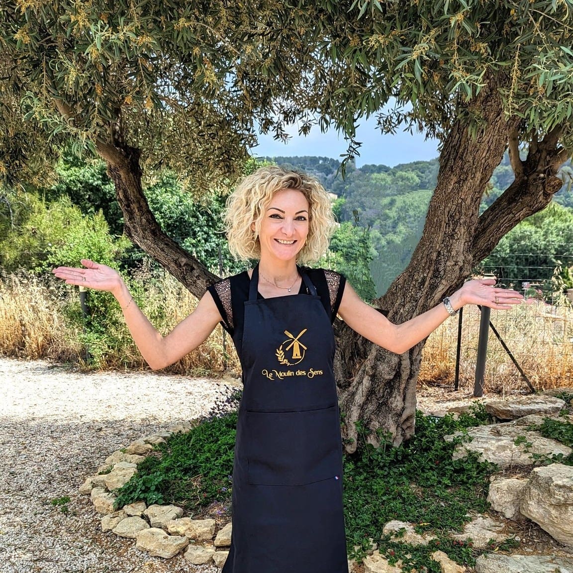 Femme souriante portant un tablier noir personnalisé "Le Moulin des Sens", posant à l’extérieur sous un olivier.