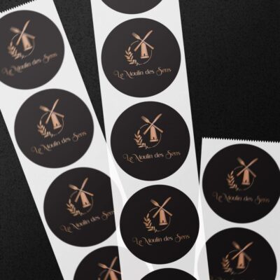 Autocollants ronds noirs avec logo doré du restaurant Le Moulin des Sens, disposés sur fond sombre.