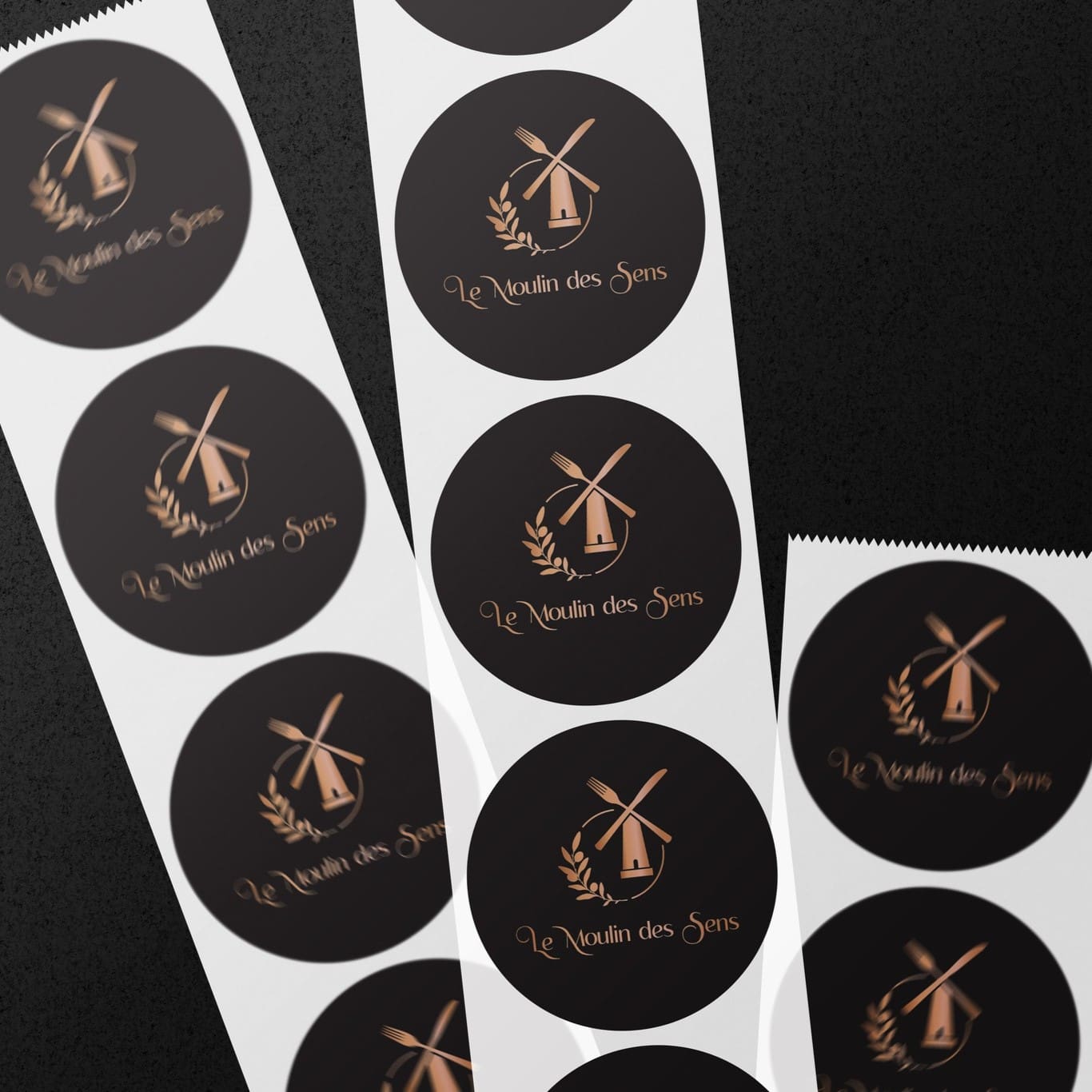 Autocollants ronds noirs avec logo doré du restaurant Le Moulin des Sens, disposés sur fond sombre.