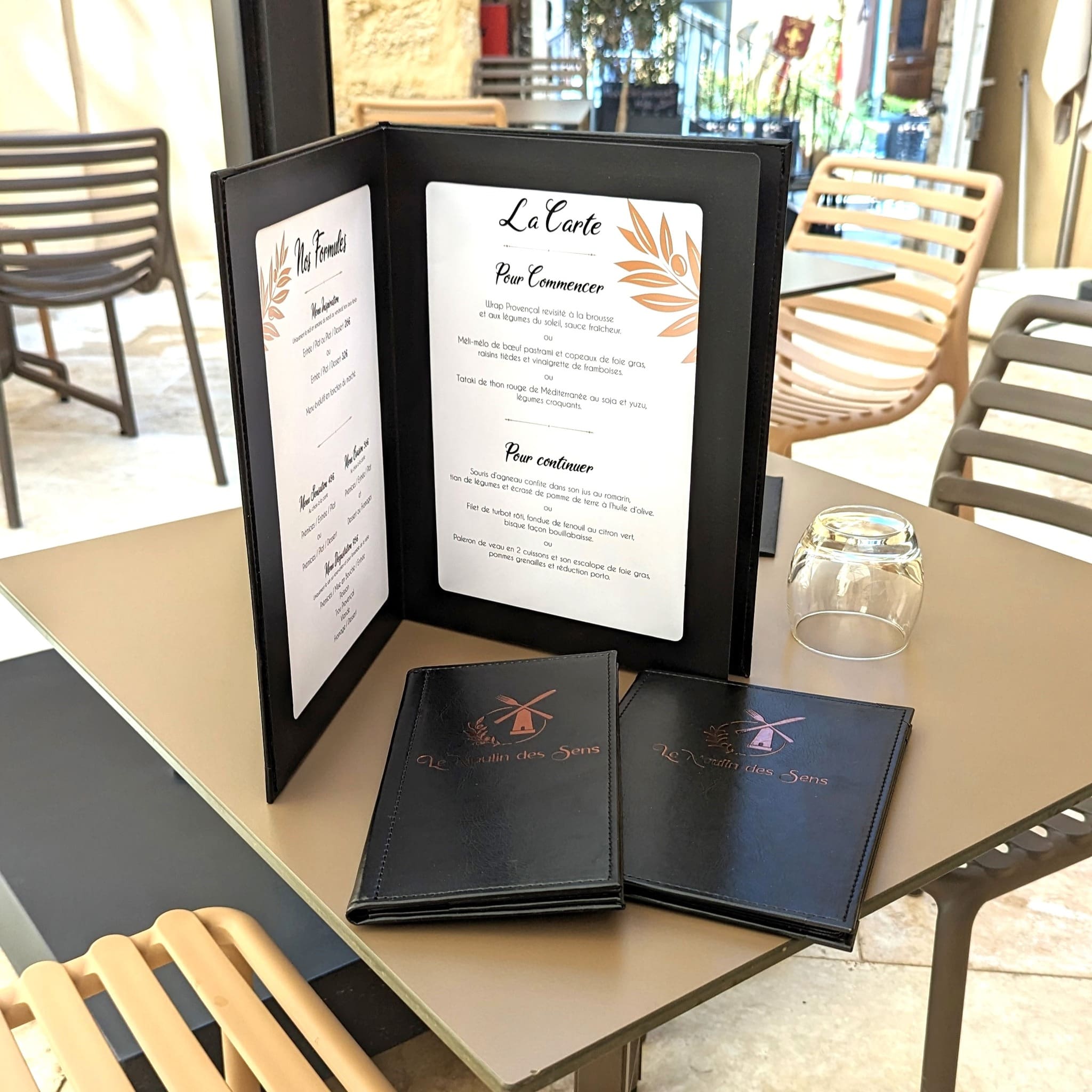 Menu du restaurant Le Moulin des Sens présenté dans une élégante chemise noire sur une table de terrasse, accompagné de deux cartes fermées et d’un verre retourné.