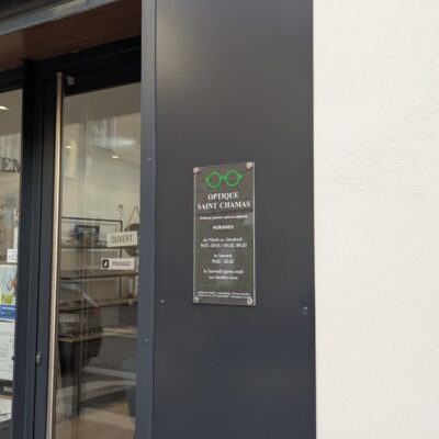 Plaque d'identification "Optique Saint Chamas" fixée à l'entrée d'une boutique, avec horaires d'ouverture.